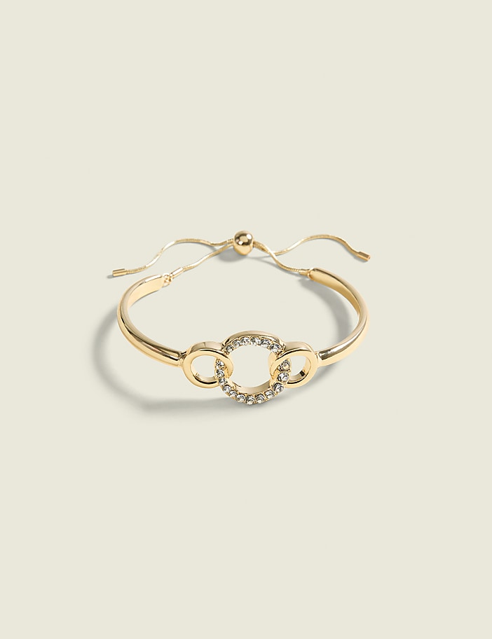 A gold bracelet with a circular, rhinestone-embellished charm and two smaller circular links rests on a pale green background.