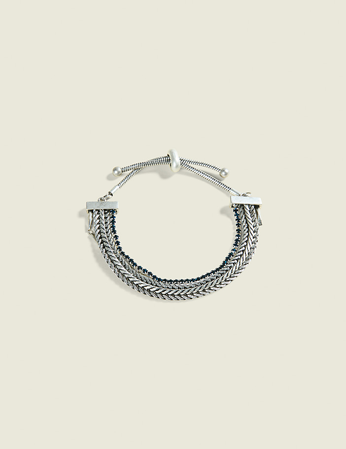 A layered silver chain bracelet with dark beads rests on a pale, textured background.