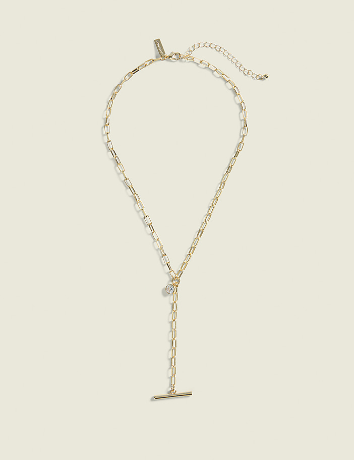A delicate gold necklace with a toggle clasp and a small charm hangs against a plain, light beige background.