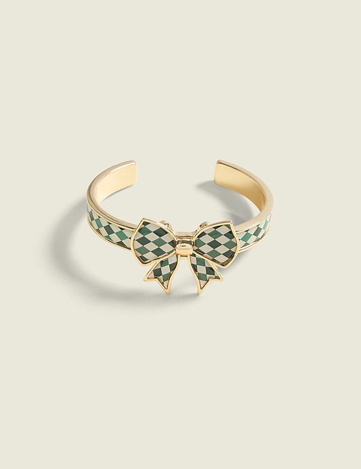 A gold ring with a green checkered bow sits on a plain, light-colored surface.