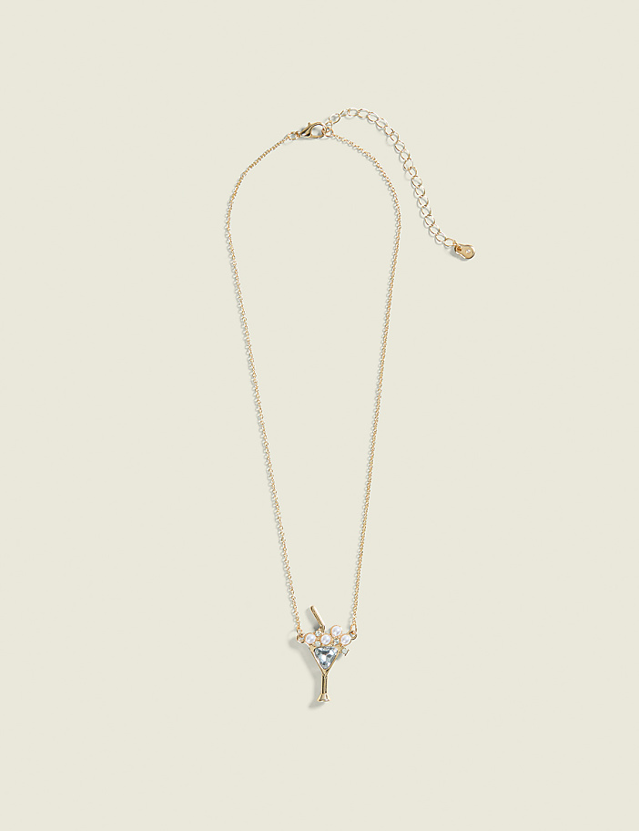 A delicate gold necklace with a martini glass charm adorned with small pearls and blue gems hangs against a plain, light beige background.