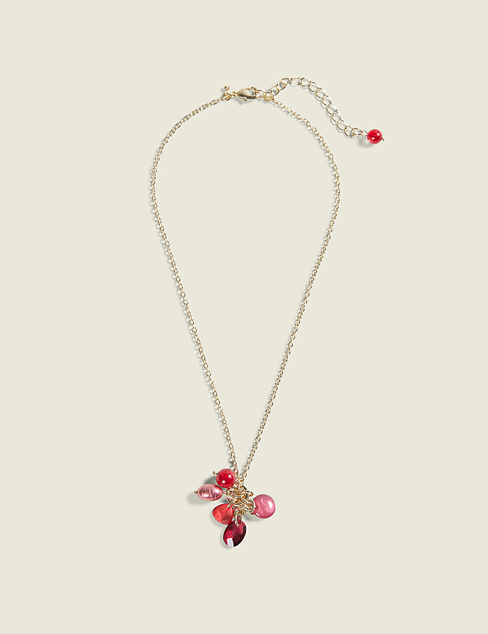 A delicate gold chain necklace with a cluster of red and pink beads hangs against a plain, light beige background.