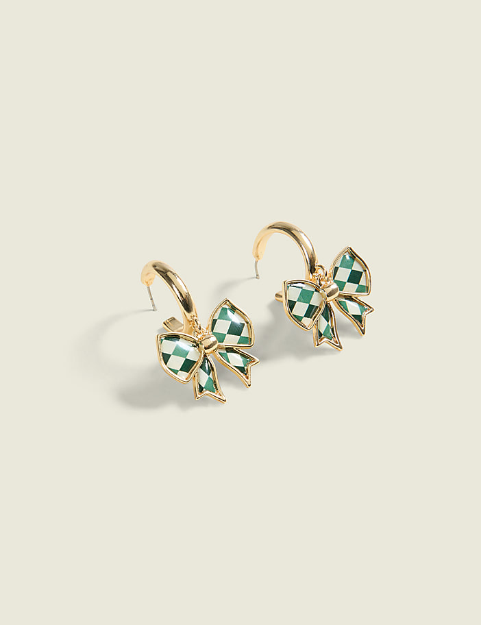 A pair of gold hoop earrings with green checkered bow charms hang against a plain, light-colored background.