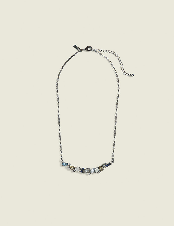 A delicate silver necklace with a curved arrangement of sparkling blue and clear stones rests on a plain, light beige background.