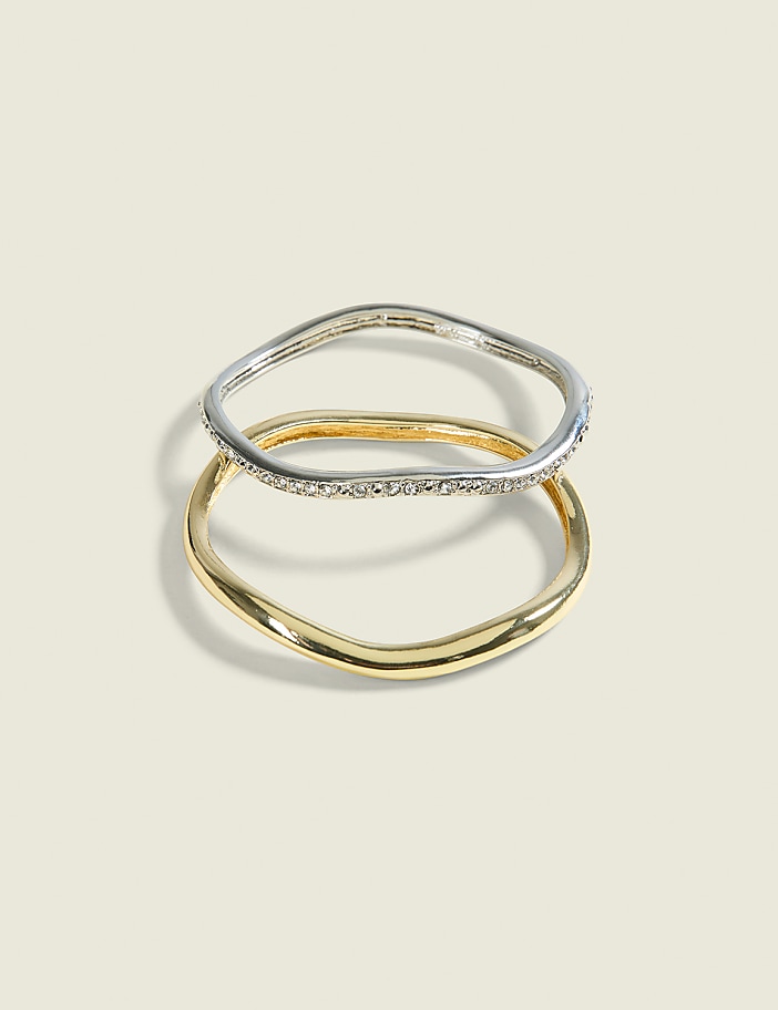 Two wavy rings, one silver with small stones and one gold, are stacked on a pale, textured background.