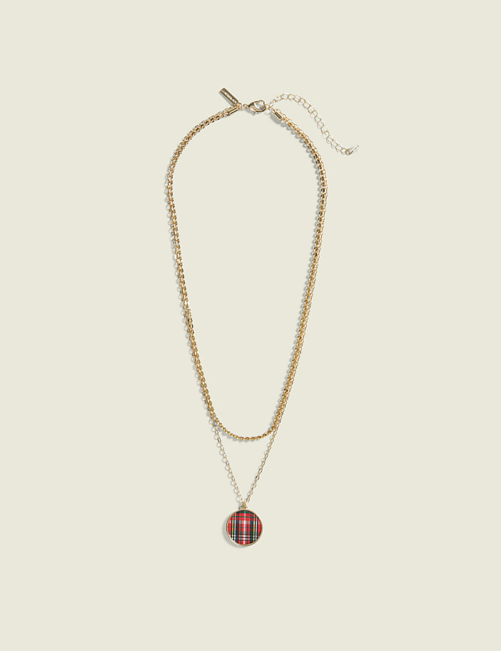 A layered gold necklace with a red and black plaid pendant hangs against a plain, light beige background.