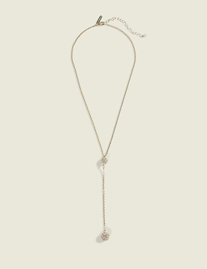 A delicate gold lariat necklace with a sparkling pendant hangs against a plain, light beige background.