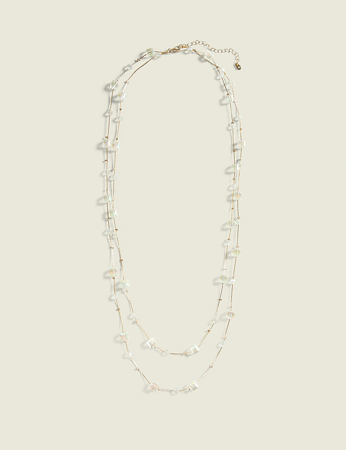 A delicate, layered necklace with small, irregular clear beads and gold accents rests against a plain, light beige background.