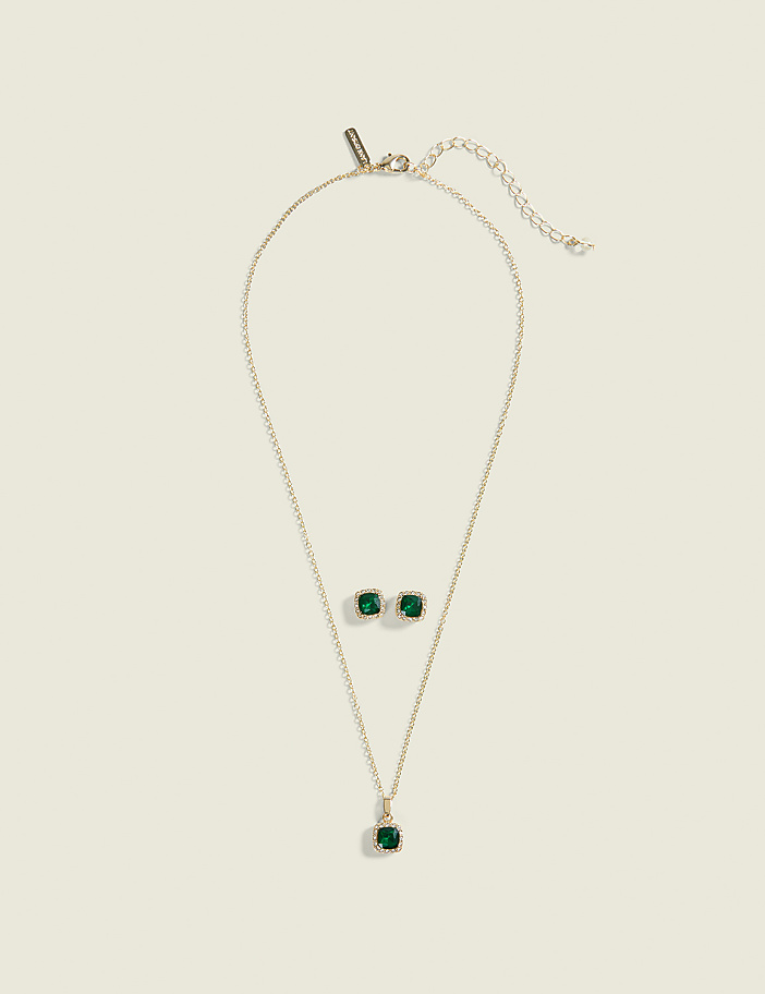 A gold necklace with a green pendant and matching square stud earrings rest on a plain, light beige background.