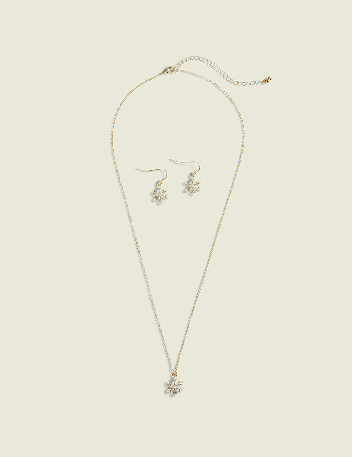 A delicate gold necklace with a snowflake pendant and matching snowflake earrings rest on a plain, light beige background.