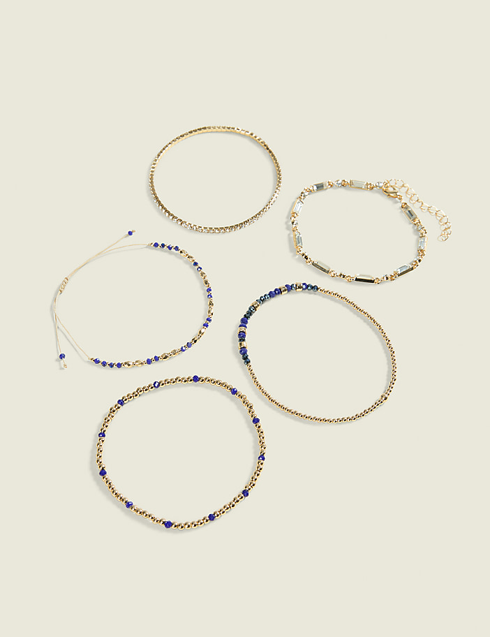 Five delicate bracelets, some adorned with blue beads and others with gold and clear beads, are arranged on a plain, light-colored background.