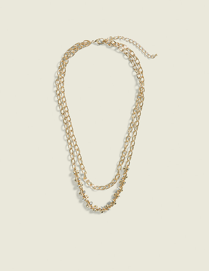 A layered gold chain necklace with small beaded accents rests on a plain, light beige background.