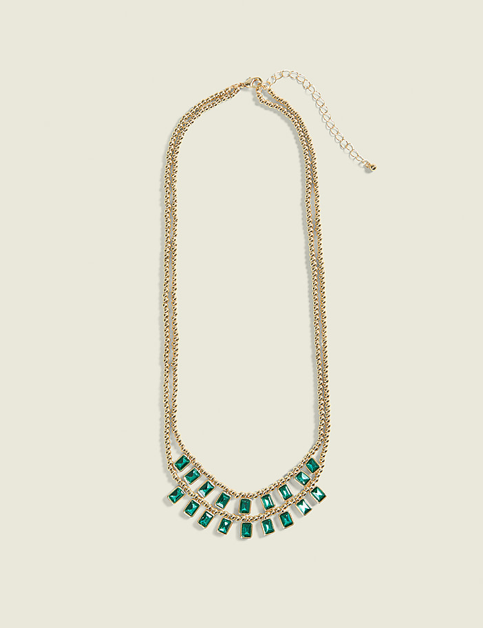 A gold necklace with emerald-colored rectangular stones hangs against a plain cream background.