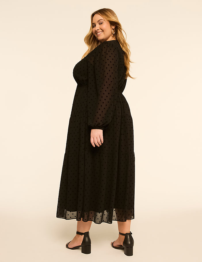 A smiling woman in a black polka-dot dress stands against a plain, light-colored background.