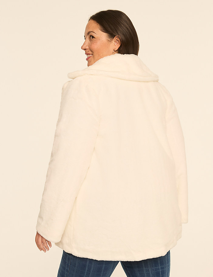 A woman in a cream-colored fuzzy jacket and blue jeans stands against a plain, light-colored background.
