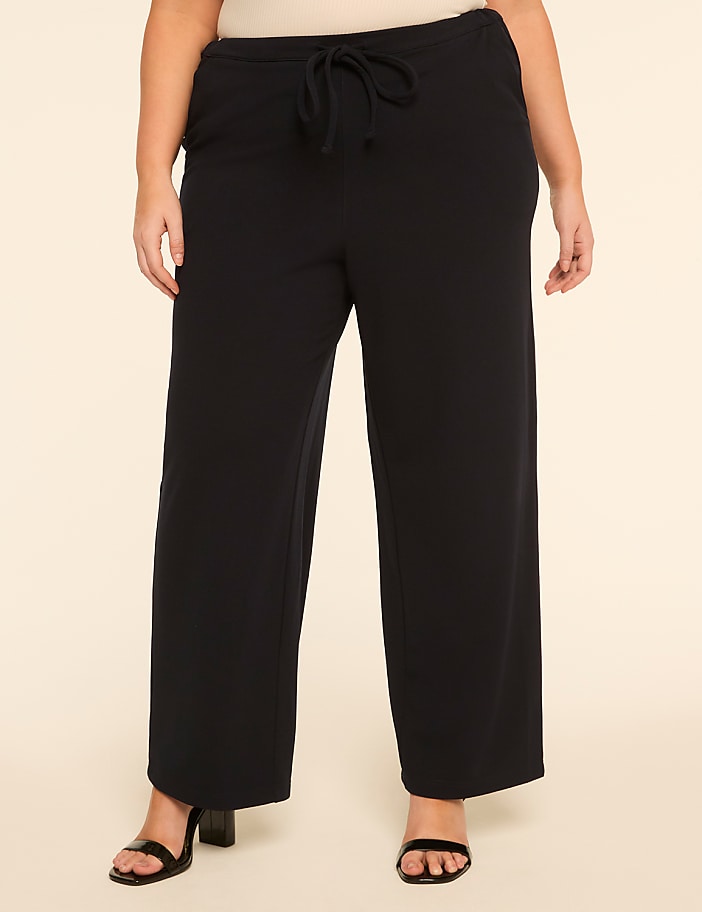 A person wearing black wide-leg pants and black heels stands against a plain, light-colored background.