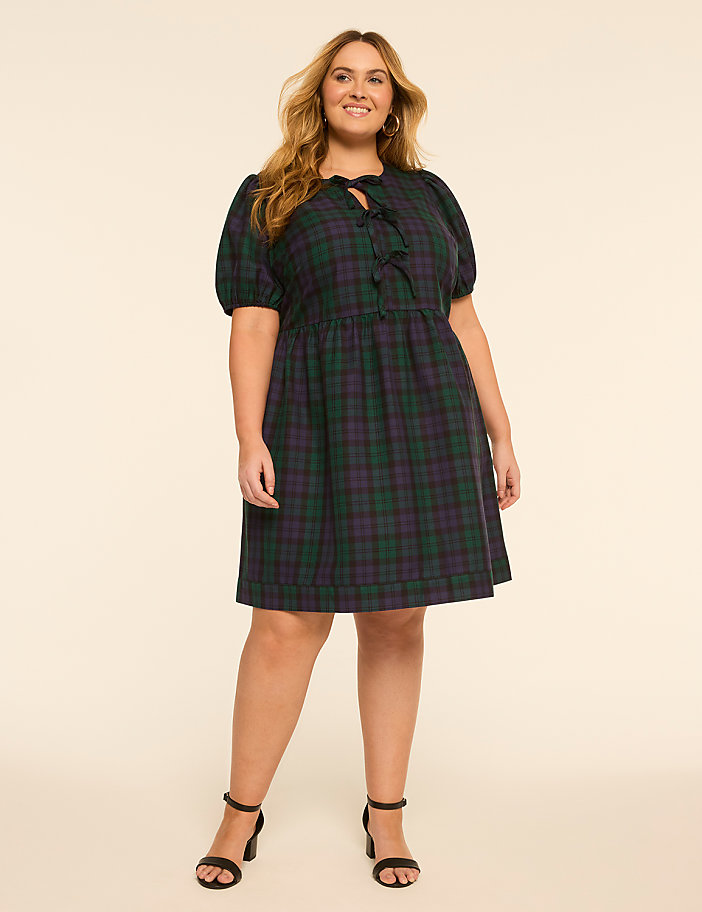 A smiling woman in a plaid dress stands against a plain, light-colored background.