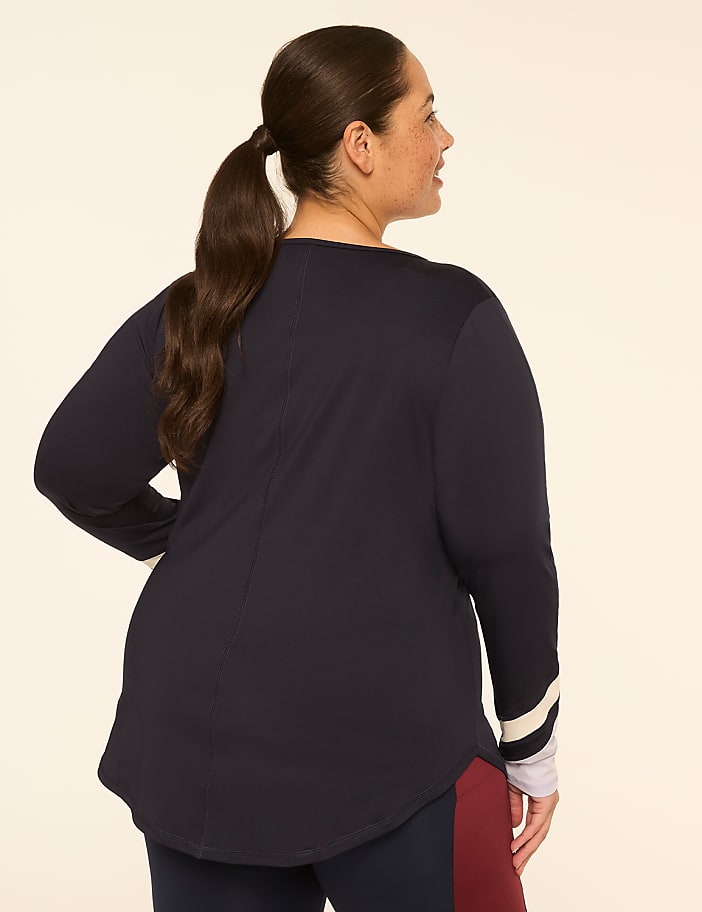 A woman with dark hair pulled into a ponytail, wearing a navy long-sleeved shirt with striped cuffs and burgundy leggings, stands against a plain, light-colored background.