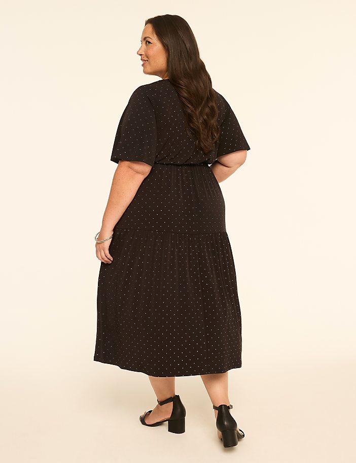 A woman in a black polka dot dress stands against a plain, light-colored background.