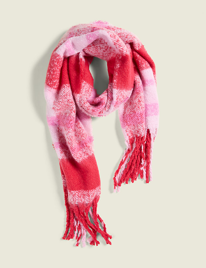 A fuzzy, plaid scarf in shades of pink and red with fringed ends rests against a plain, light beige background.