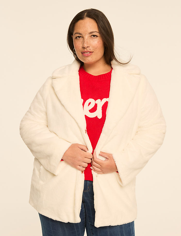 A woman in a white faux fur coat and red sweater stands against a plain, light-colored background.