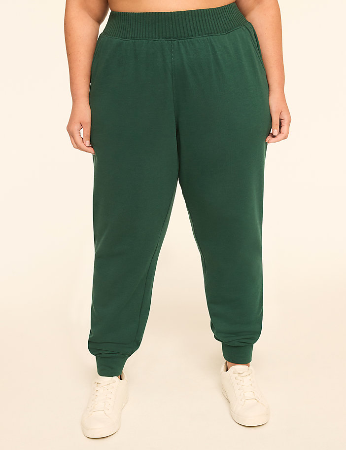 A person wearing dark green sweatpants and white sneakers stands against a plain, light-colored background.