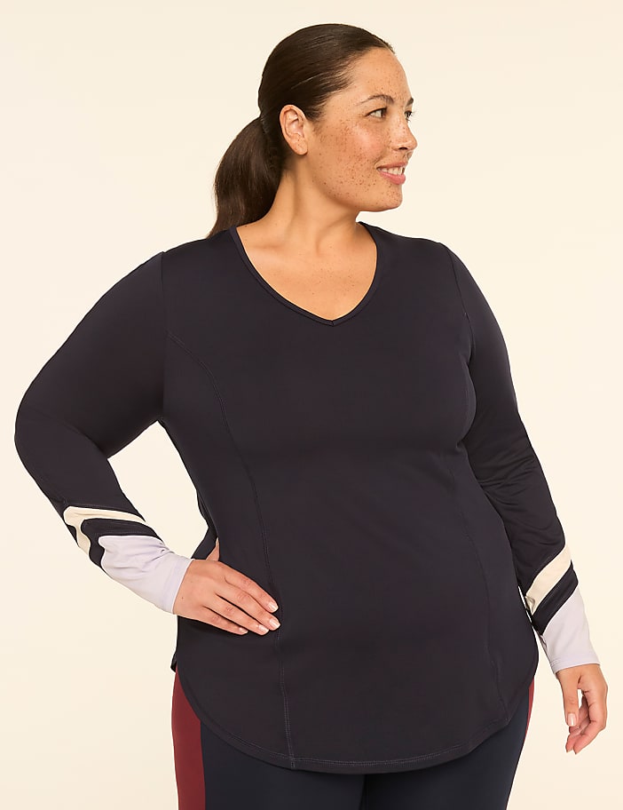A woman with dark hair pulled back in a ponytail poses with her hand on her hip, wearing a navy v-neck shirt with striped cuffs, against a plain, light-colored background.