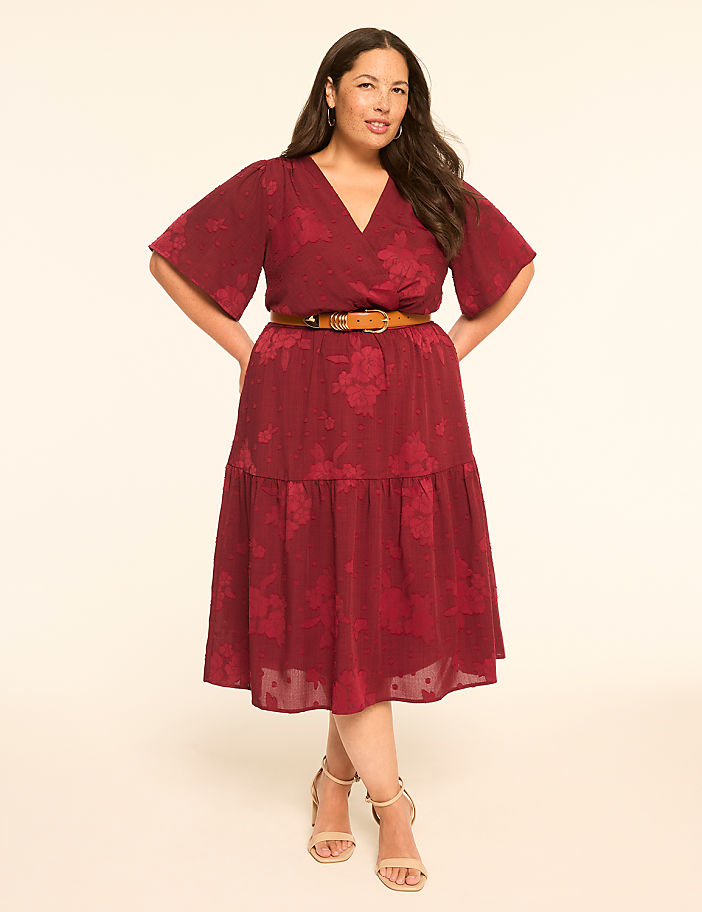 A woman in a burgundy floral dress and tan heels stands against a plain, light background.