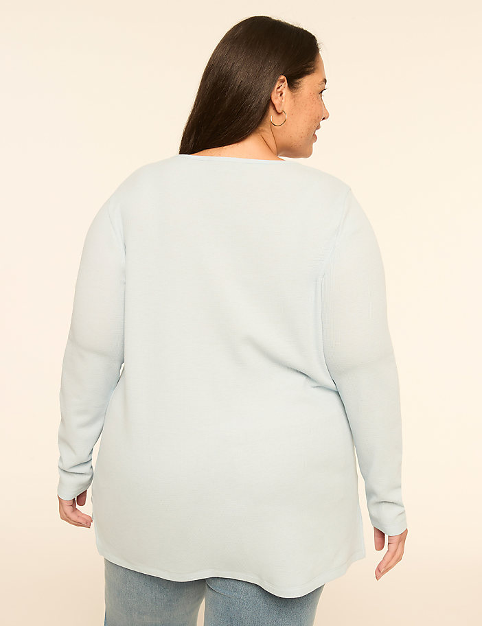 A woman in a light blue sweater and jeans stands against a plain, light-colored background.