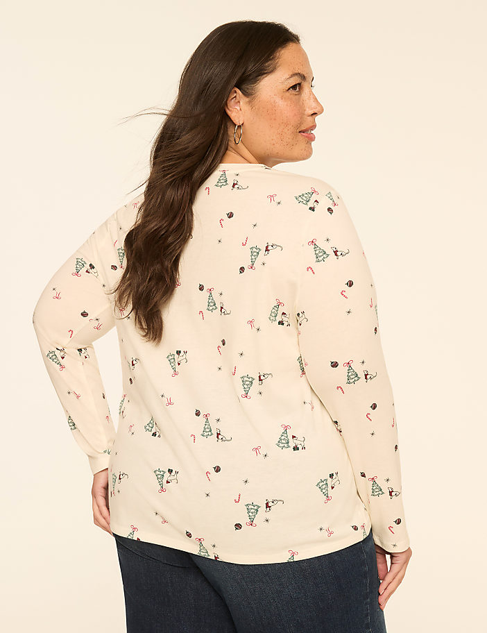 A woman in a cream-colored long-sleeved shirt with a festive Christmas tree and candy cane pattern stands against a plain, light-colored background.