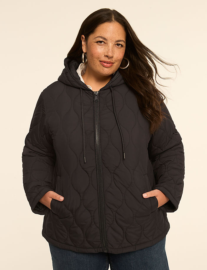 A woman in a black quilted hooded jacket stands against a plain, light-colored background.