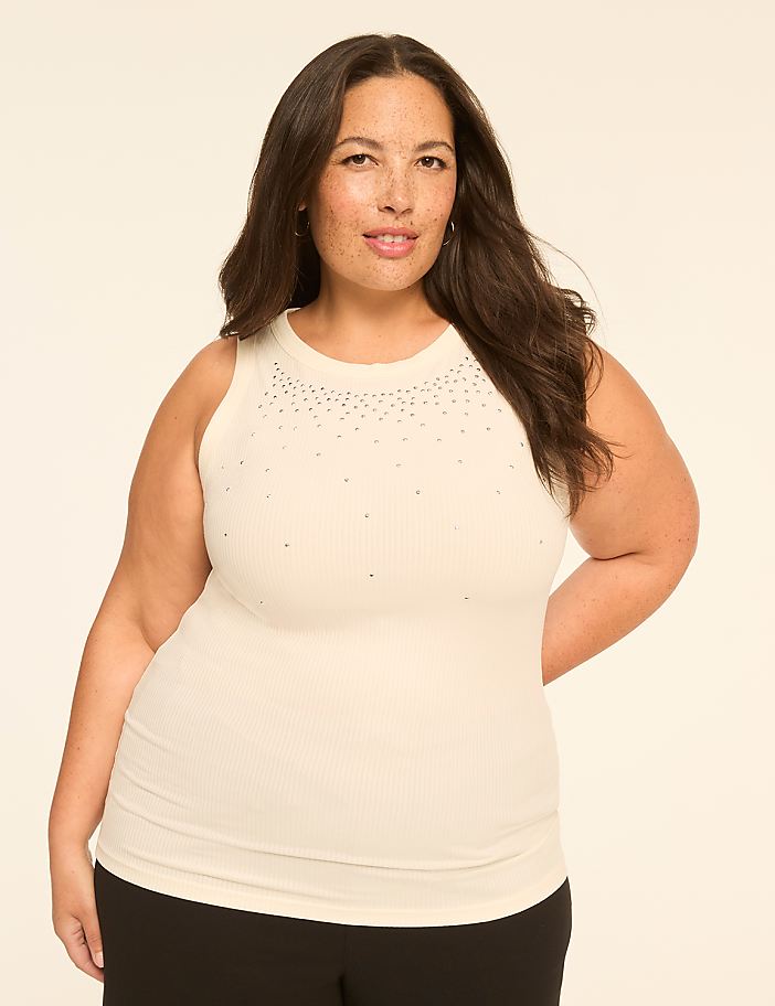 A woman in a cream-colored, sleeveless, ribbed top with subtle embellishments on the neckline stands against a plain, light-colored background.