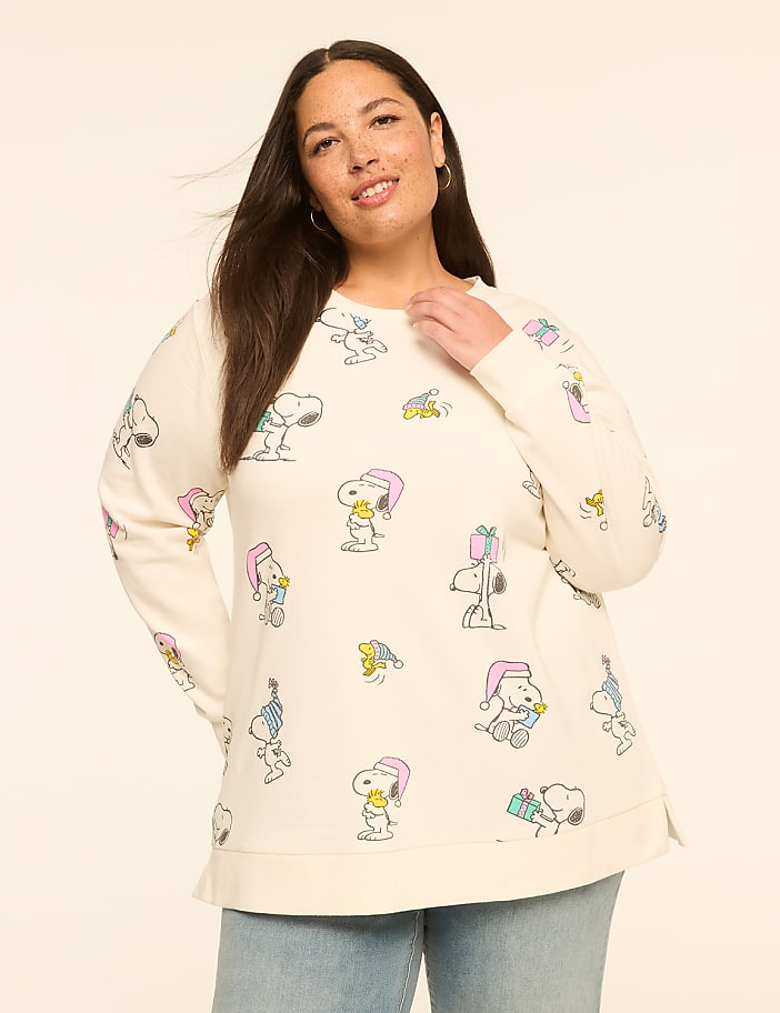 A woman in a cream-colored sweatshirt with a Snoopy and Woodstock pattern stands against a plain, light beige background.