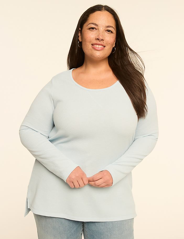 A smiling woman with long dark hair and a light blue long-sleeved shirt stands against a plain, light-colored background.