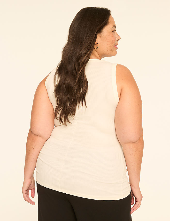 A woman with long, dark hair wearing a cream-colored tank top stands against a plain, light-colored background.