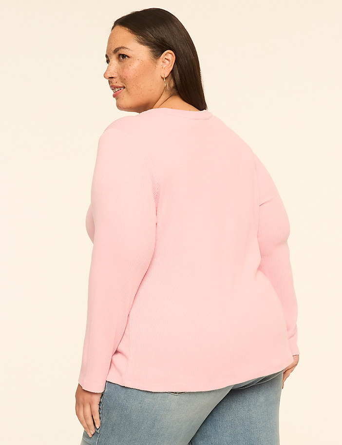 A woman in a pink waffle-knit shirt and blue jeans stands against a plain, light-colored background.