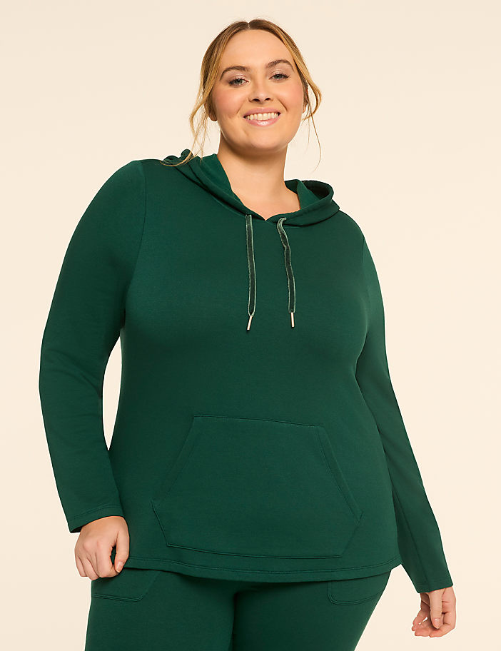 A smiling woman in a green hoodie and matching pants stands against a plain, light-colored background.