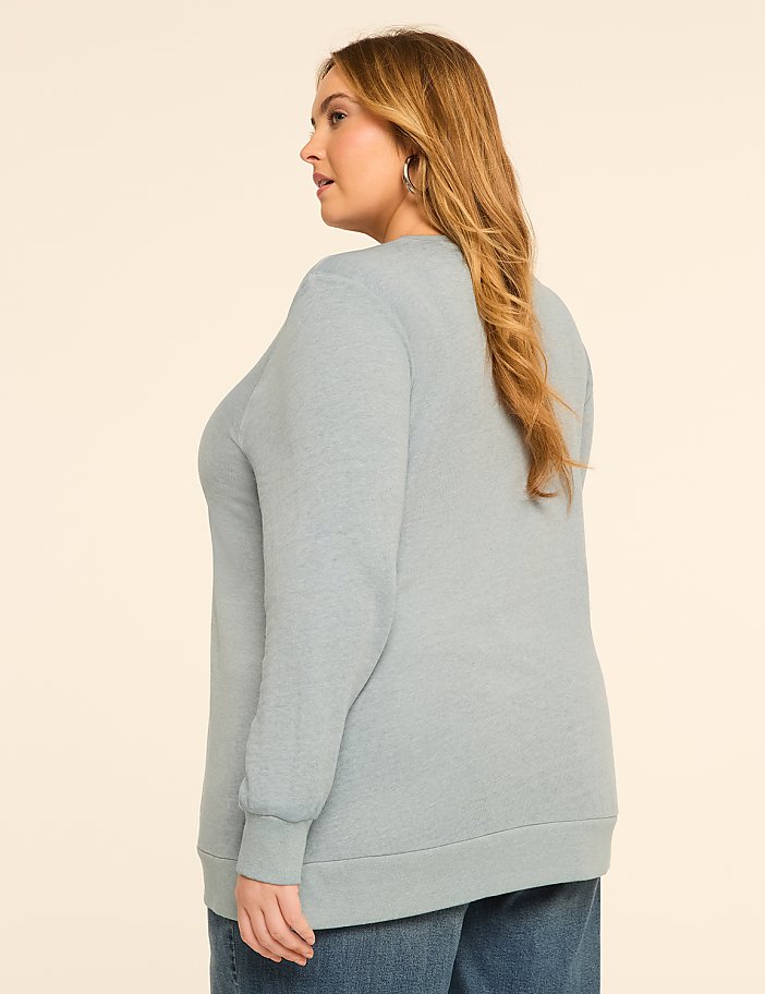A woman in a light blue sweatshirt and jeans stands against a plain, light-colored background.