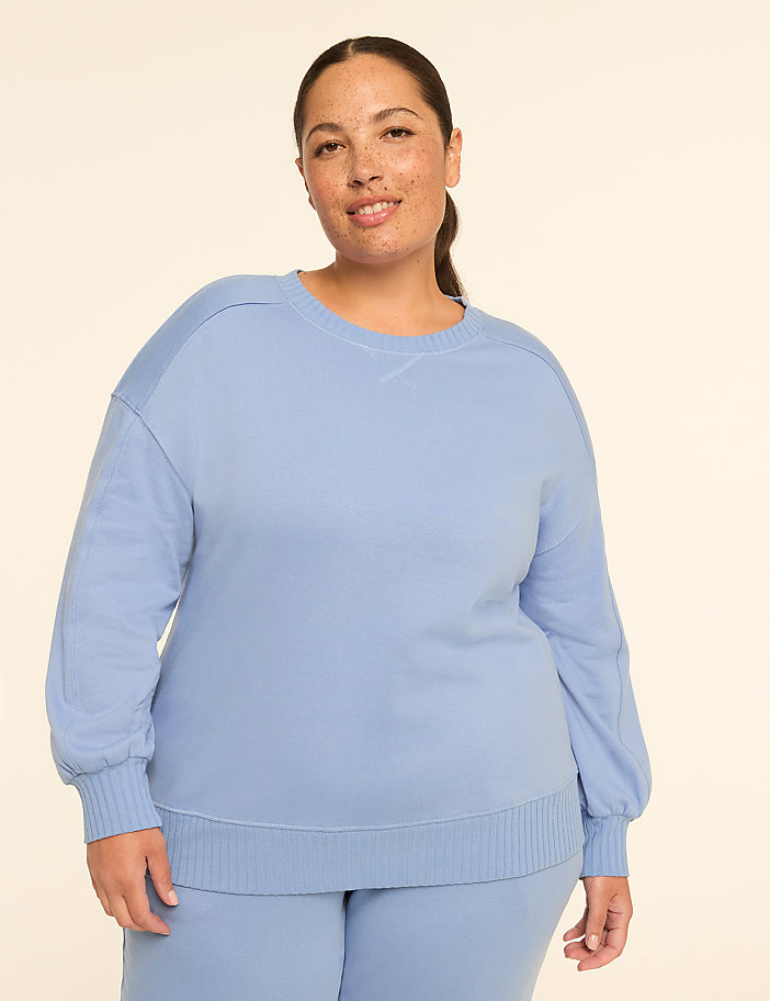 A smiling woman with freckles in a light blue sweatshirt and matching pants stands against a plain, light background.