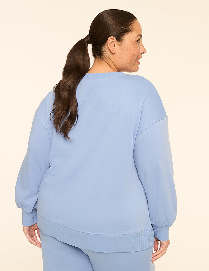 A woman in a light blue sweatshirt and pants stands against a plain, light-colored background.