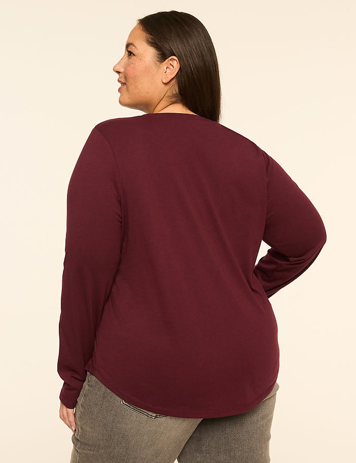 A woman in a maroon long-sleeved shirt and grey jeans stands against a plain, light-colored background.