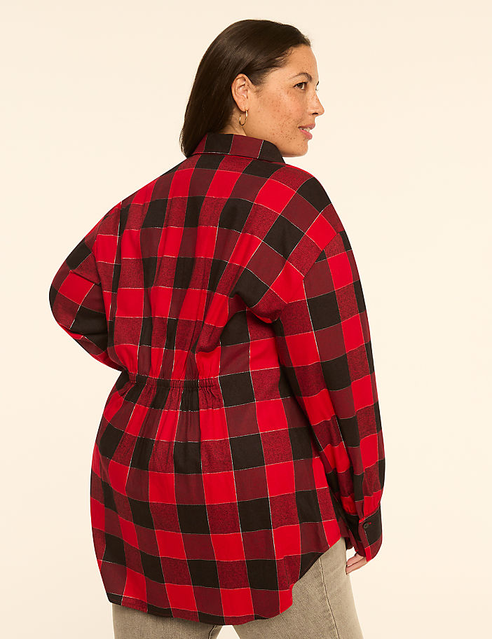 A woman in a red and black plaid shirt with a cinched waist stands against a plain, light-colored background.