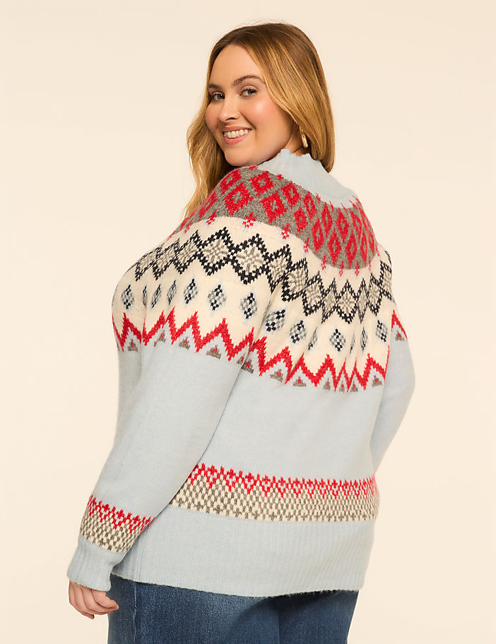 A woman in a light blue sweater with a fair isle pattern smiles over her shoulder against a plain, light-colored background.