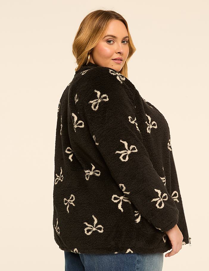 A woman in a black fuzzy jacket with a bow pattern stands against a plain, light-colored background.