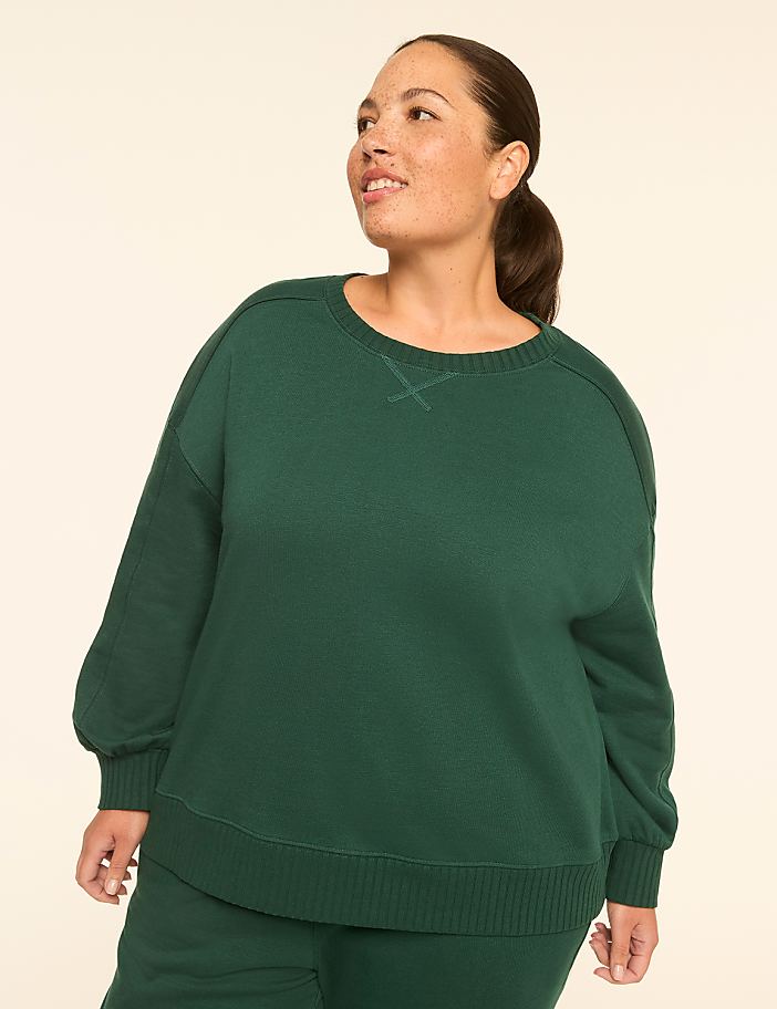 A woman in a green sweatshirt smiles as she looks to the side against a plain, light-colored background.