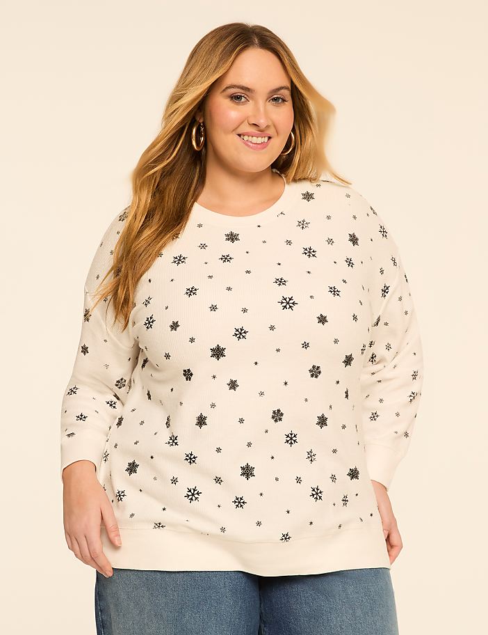 A smiling woman with blonde hair and hoop earrings wears a cream-colored sweatshirt with a snowflake pattern, standing against a plain, light-colored background.