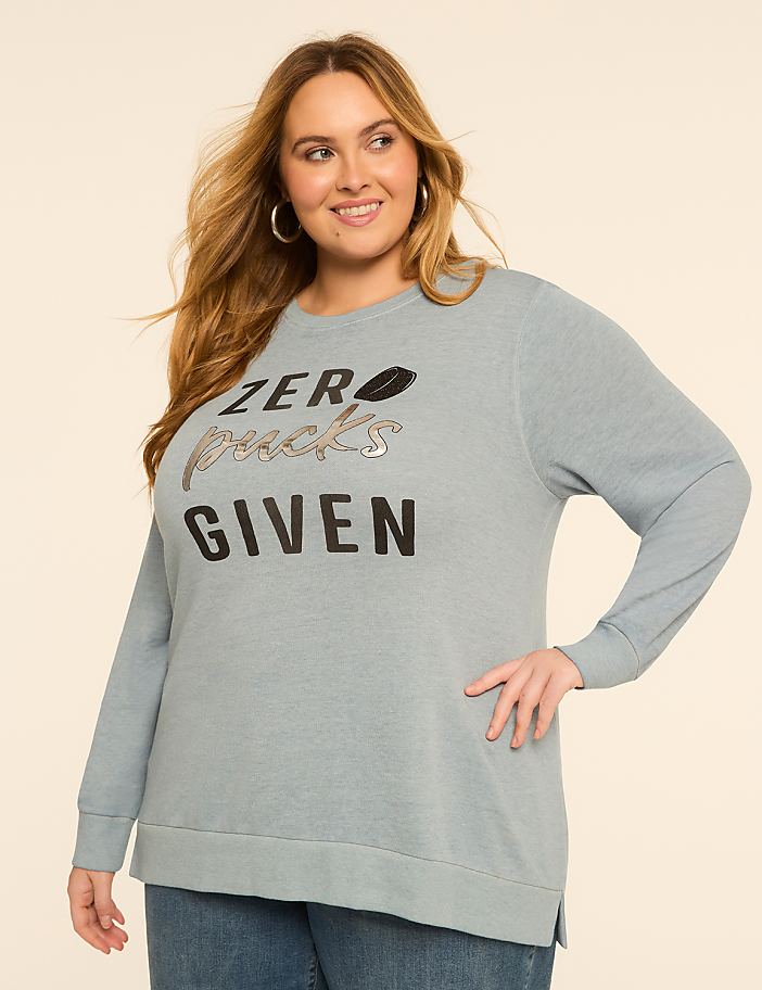 A smiling woman in a light blue sweatshirt with "ZERO PUCKS GIVEN" printed on it stands against a plain, light-colored background.