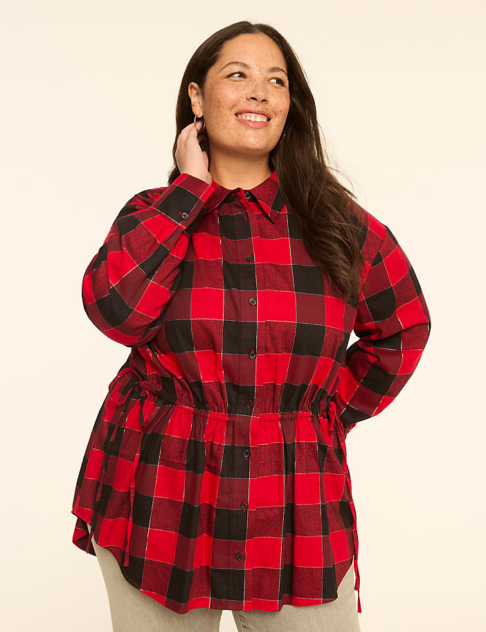 A woman in a red and black plaid shirt stands against a plain, light-colored background.
