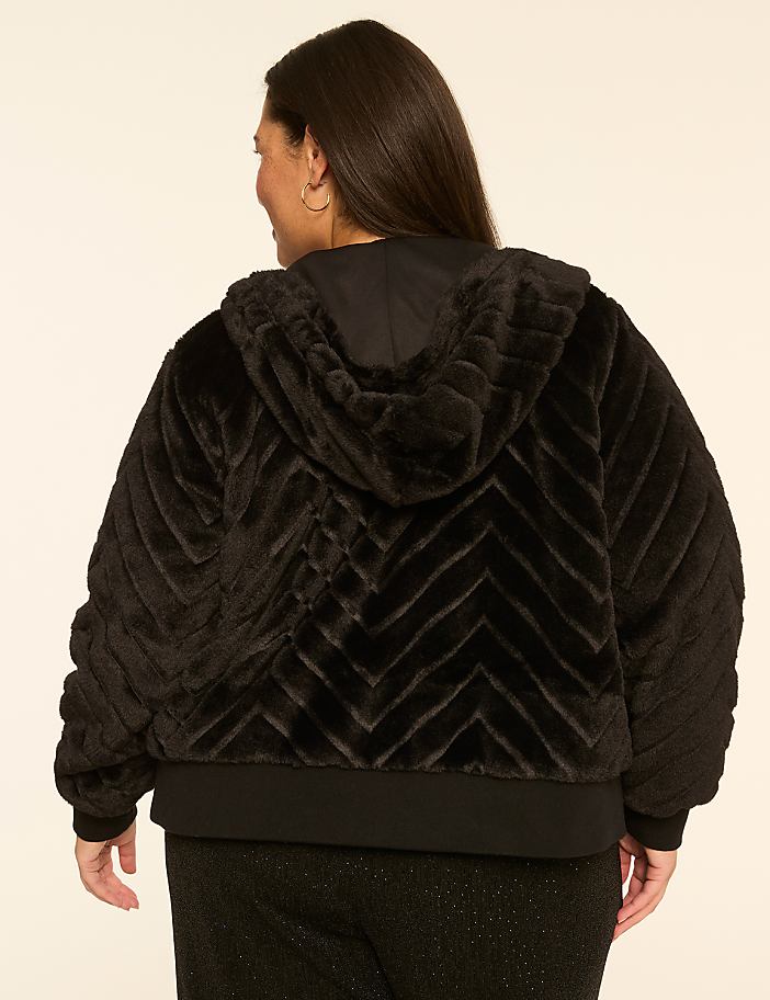 A woman in a black faux fur chevron-patterned hooded jacket stands against a plain, light-colored background.