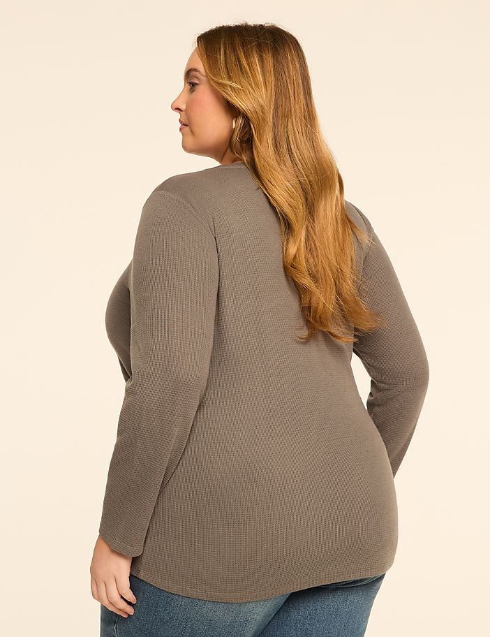 A woman with long, brown hair, wearing a grey waffle-knit top and blue jeans, stands against a plain, light-colored background.
